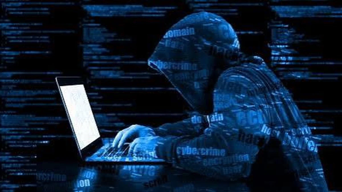 Una persona encapuchada escribe en una notebook frente a una pantalla azul con textos sobre ciberdelito