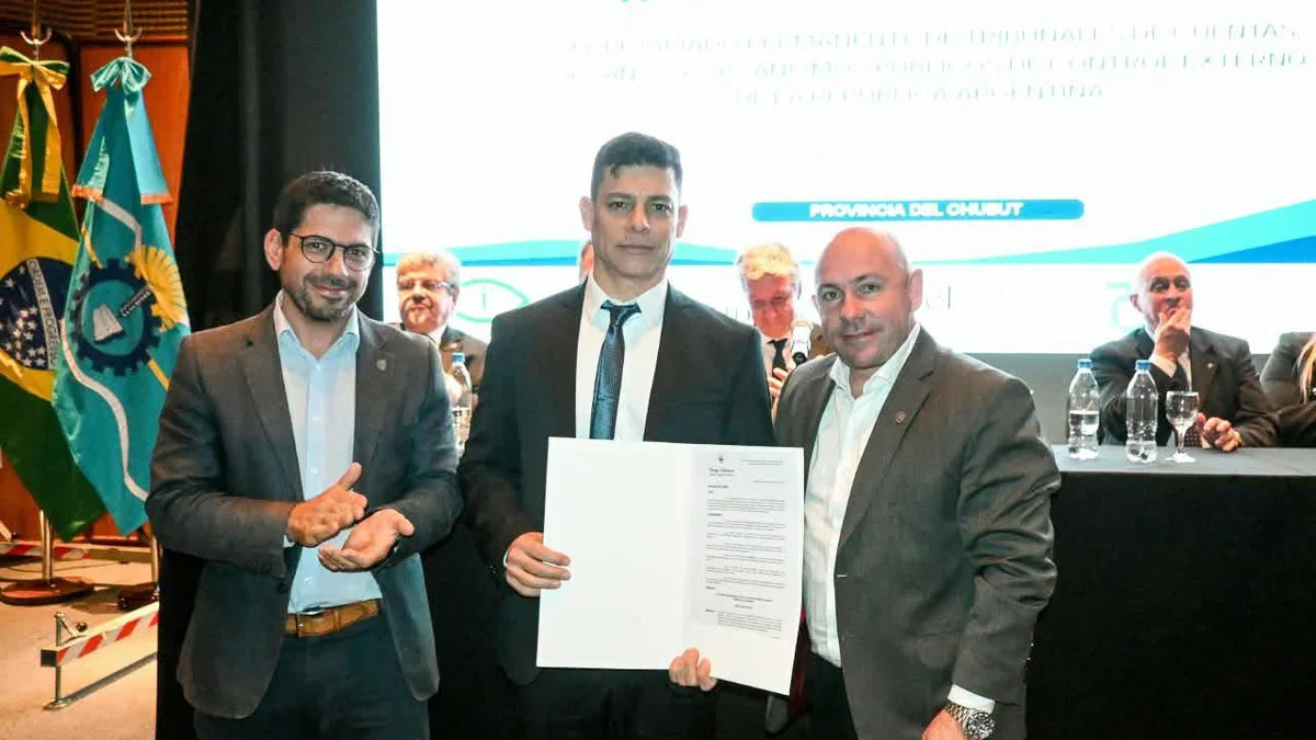 Unos tres hombres de traje posan en un escenario; el del centro sostiene una hoja con texto.
