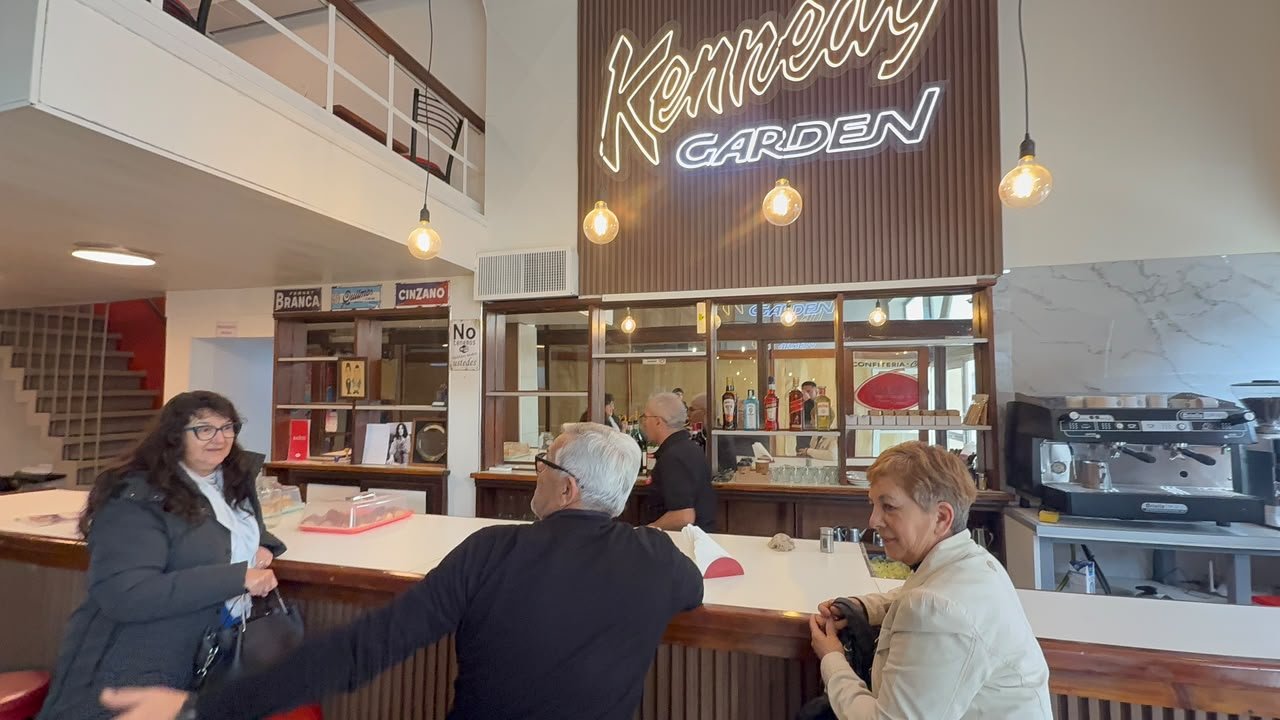 Dos personas apoyadas en la barra del Café Kennedy, bajo un cartel luminoso con el nombre del local