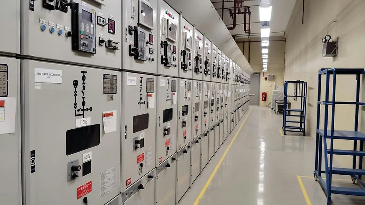 Una fila de tableros eléctricos metálicos grises con perillas, etiquetas y llaves, junto a un pasillo de piso liso
