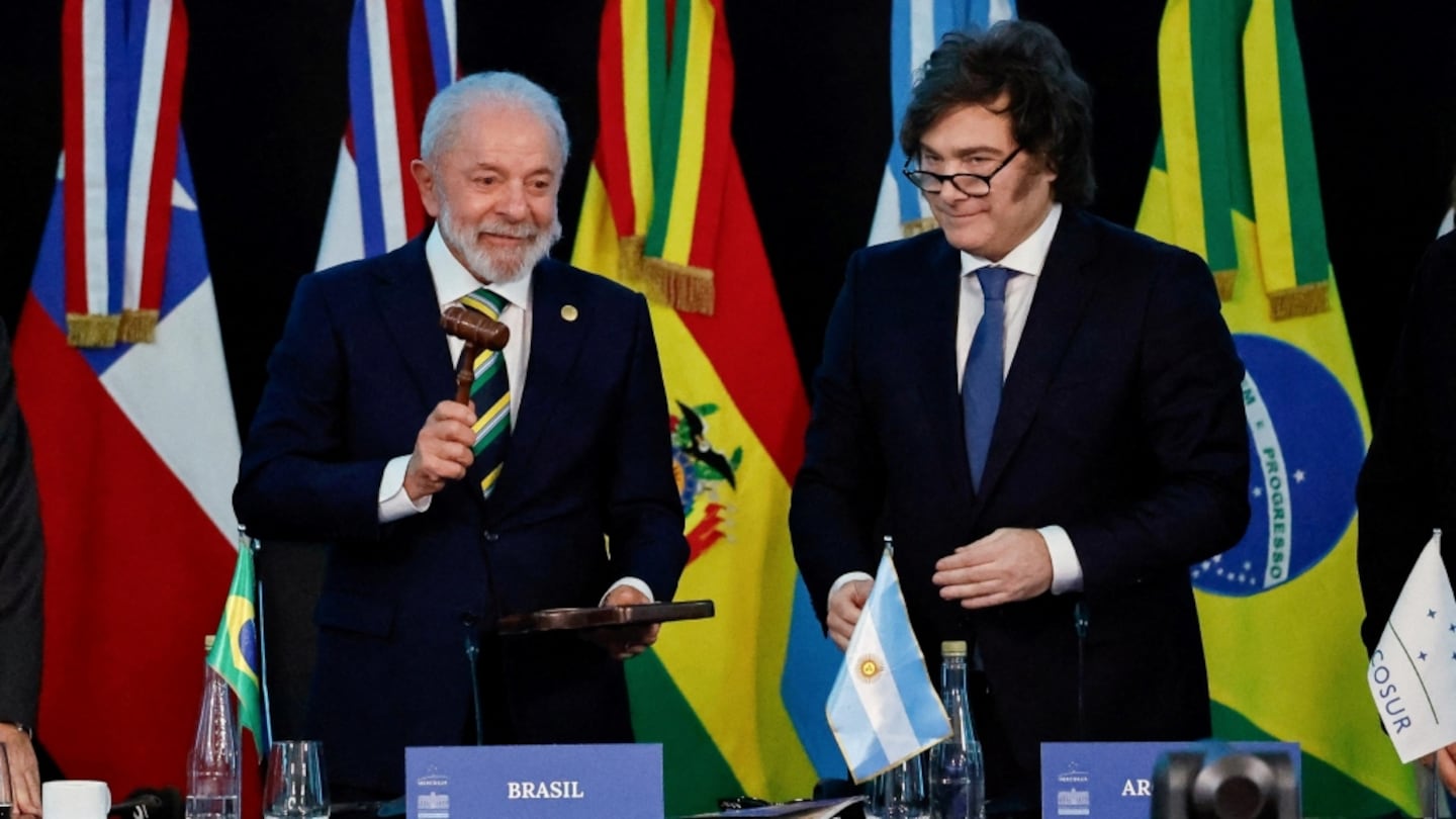 Dos hombres de traje en una mesa con banderas de Brasil y Argentina; uno sostiene un mazo de madera.