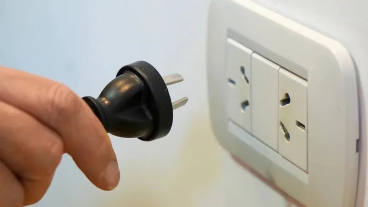 Una mano sostiene un enchufe negro frente a un tomacorriente de pared blanco