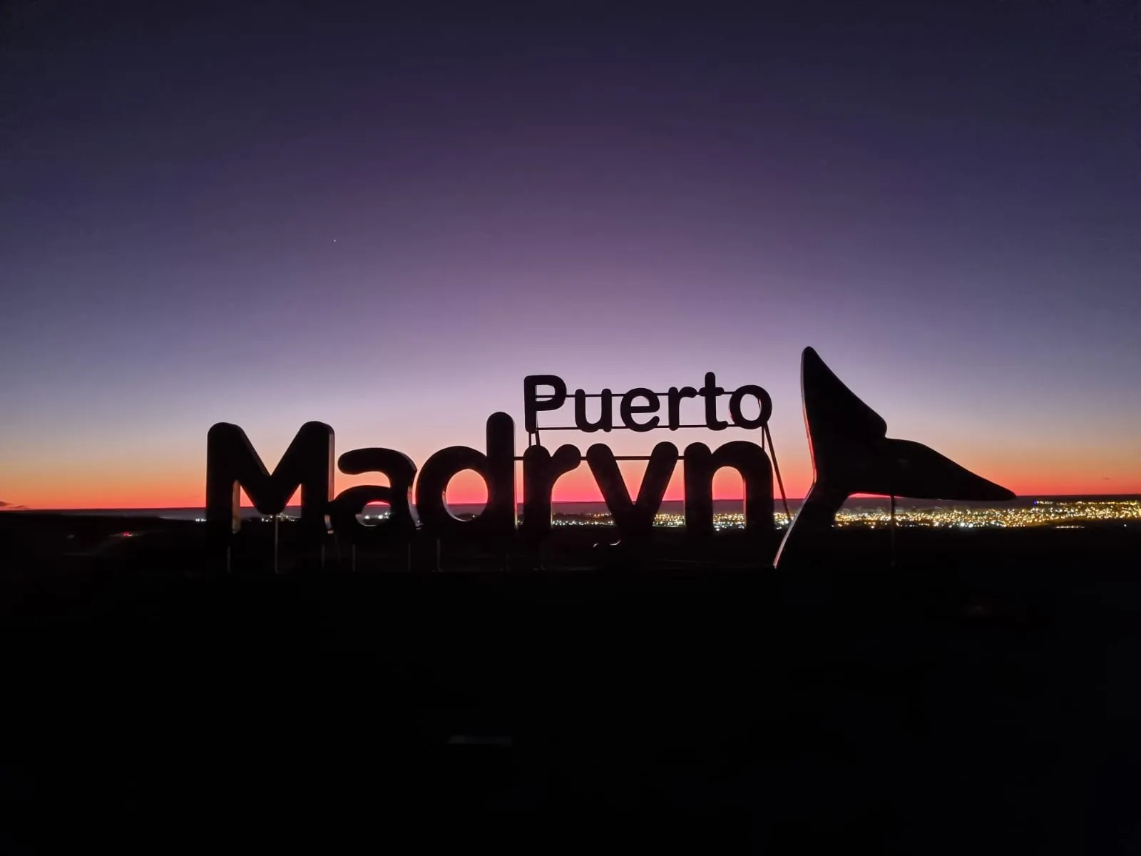 Un cartel con las palabras Puerto Madryn recortado contra el atardecer, con la ciudad iluminada detrás.