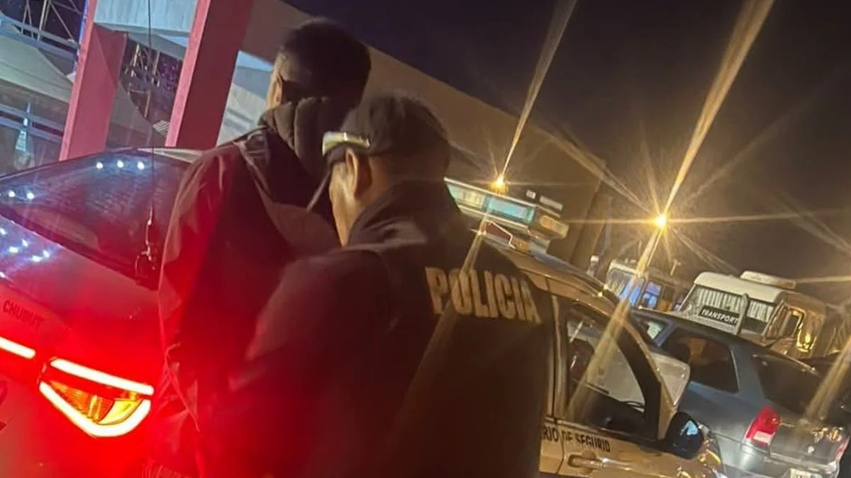 Un policía con uniforme oscuro acompaña a un hombre encapuchado junto a un patrullero, de noche.