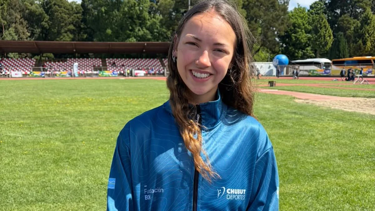 Una joven sonriente con campera azul posa en un campo de atletismo con gradas al fondo.