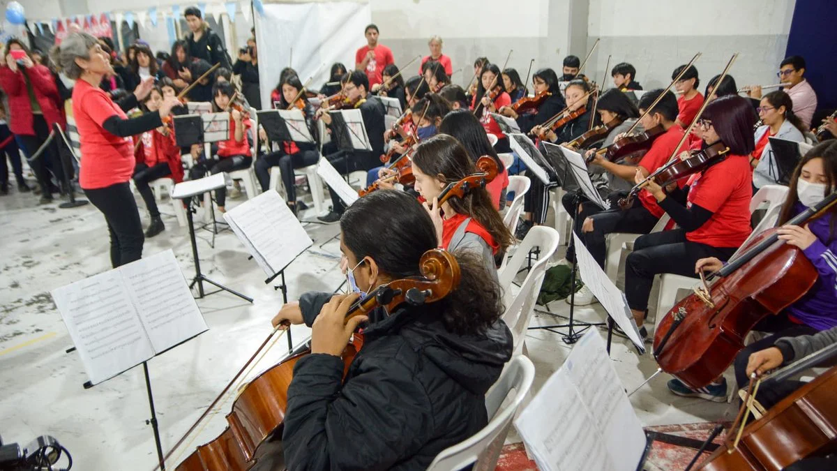 Una directora de pelo canoso y remera roja dirige a jóvenes músicos que tocan violines y violonchelos frente a sus atriles