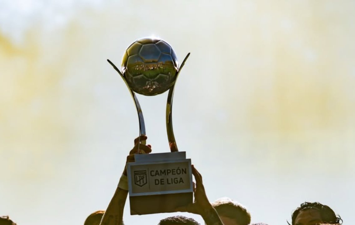 Un trofeo dorado con una pelota arriba es sostenido en alto por manos, con una placa que dice Campeón de Liga
