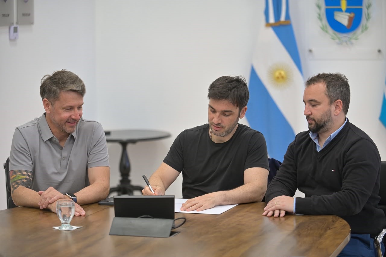 Unos tres hombres sentados a una mesa de madera, uno firma un papel frente a una bandera argentina