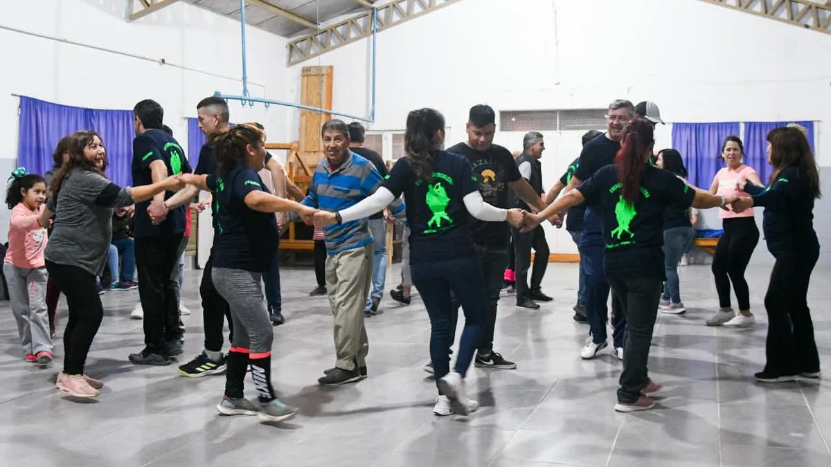 Un grupo de personas baila en círculo, tomadas de las manos, en un salón con piso gris y cortinas violetas.
