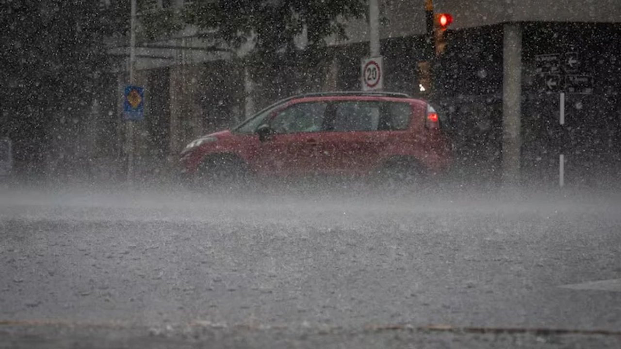 Un auto rojo avanza por una calle cubierta de lluvia intensa, con el agua salpicando desde el pavimento.