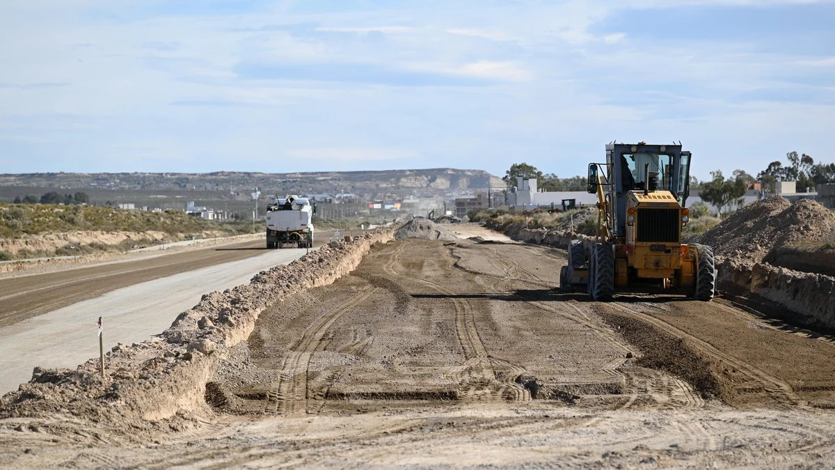 Una motoniveladora amarilla nivela el suelo de una obra vial mientras un camión cisterna avanza por la calzada