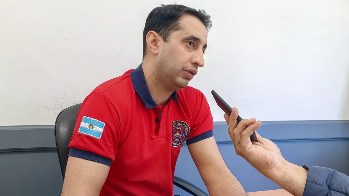 Un bombero con remera roja y bordado de bomberos voluntarios habla frente a un micrófono en un despacho