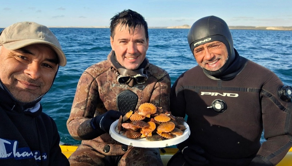 Unos hombres con trajes de buceo posan sobre una embarcación con un plato de vieiras y el mar de fondo.