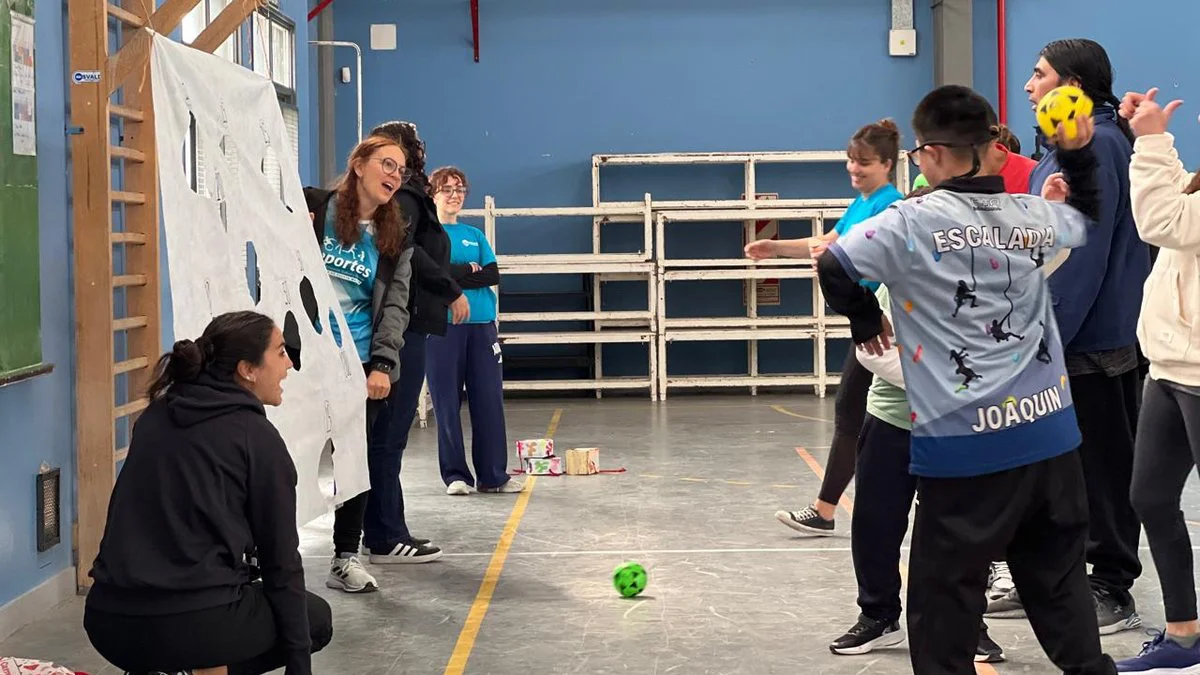 Unos jóvenes juegan en un gimnasio con una pelota verde en el piso y un panel blanco con agujeros.