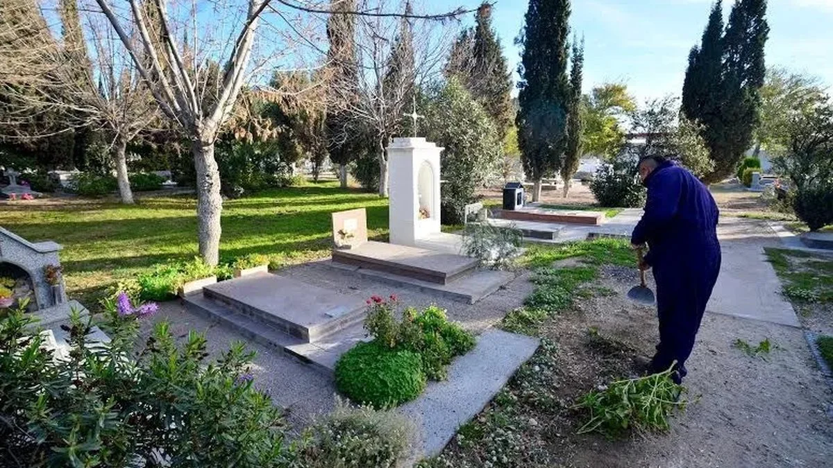 Un hombre con overol azul rastrilla la tierra junto a una tumba con un monumento blanco en el cementerio.