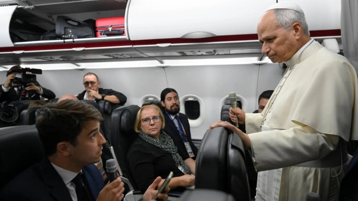 Un hombre vestido de blanco sostiene un micrófono dentro de un avión, frente a periodistas sentados en sus asientos.