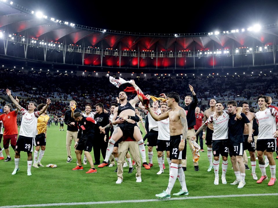 Unos jugadores con camisetas blancas y rayas rojas celebran sobre el césped de un estadio iluminado de noche.