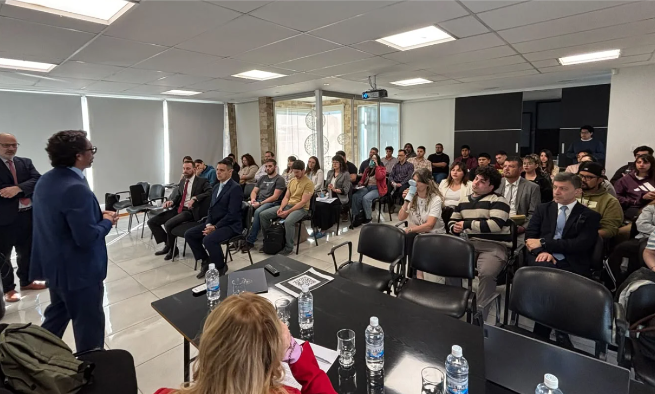 Un hombre de traje azul habla de pie frente a un grupo de personas sentadas en una sala de reuniones