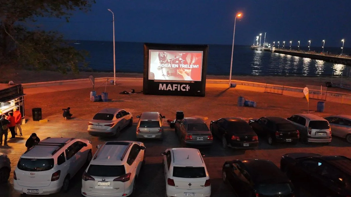 Un cine al aire libre con autos estacionados frente al mar de noche y una pantalla inflable proyectando una película.