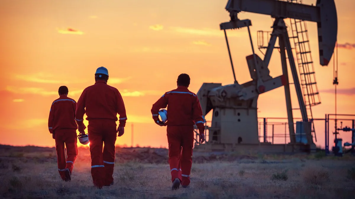 Unos trabajadores con overoles rojos y cascos caminan hacia una bomba petrolera al atardecer
