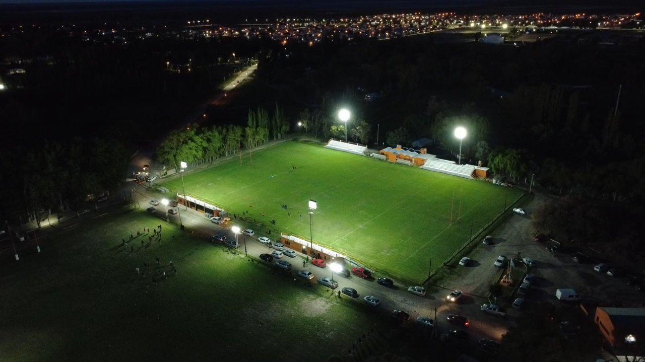 Vista aérea nocturna de una cancha de rugby iluminada por cuatro torres de luz, con autos estacionados al costado.