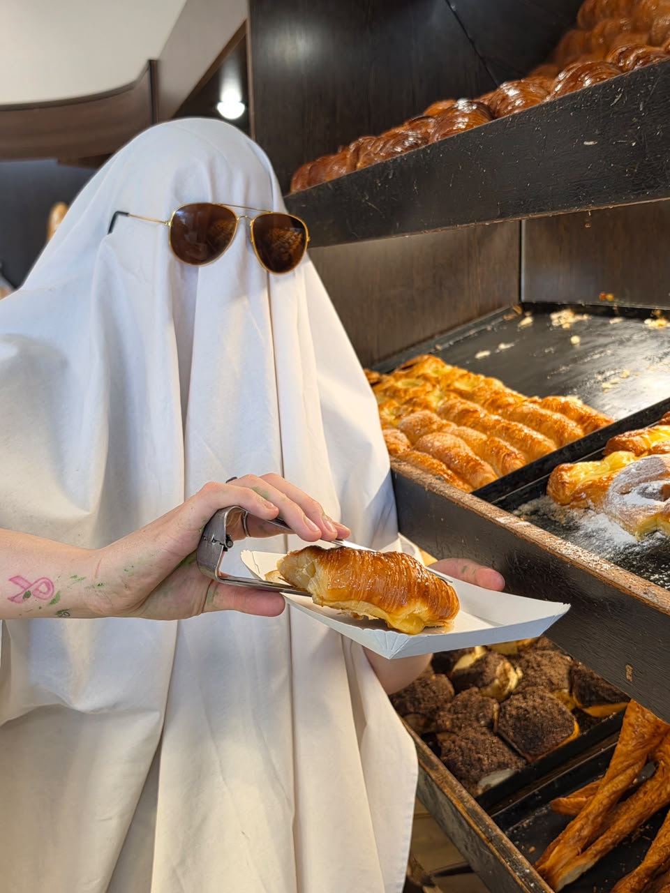 Una persona disfrazada de fantasma con anteojos de sol toma un croissant con pinzas en una panadería.