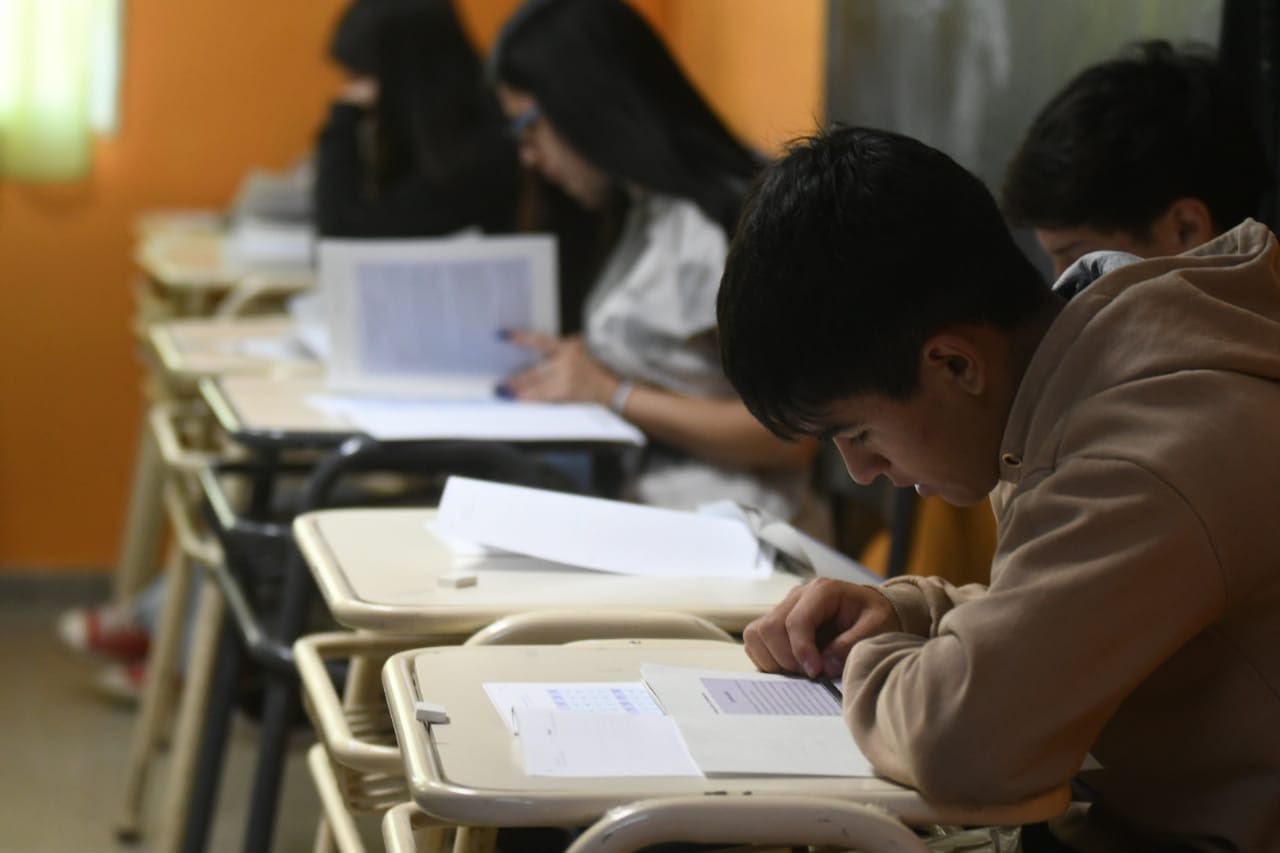 Un adolescente con buzo marrón escribe en un pupitre de un aula, con compañeros al fondo.