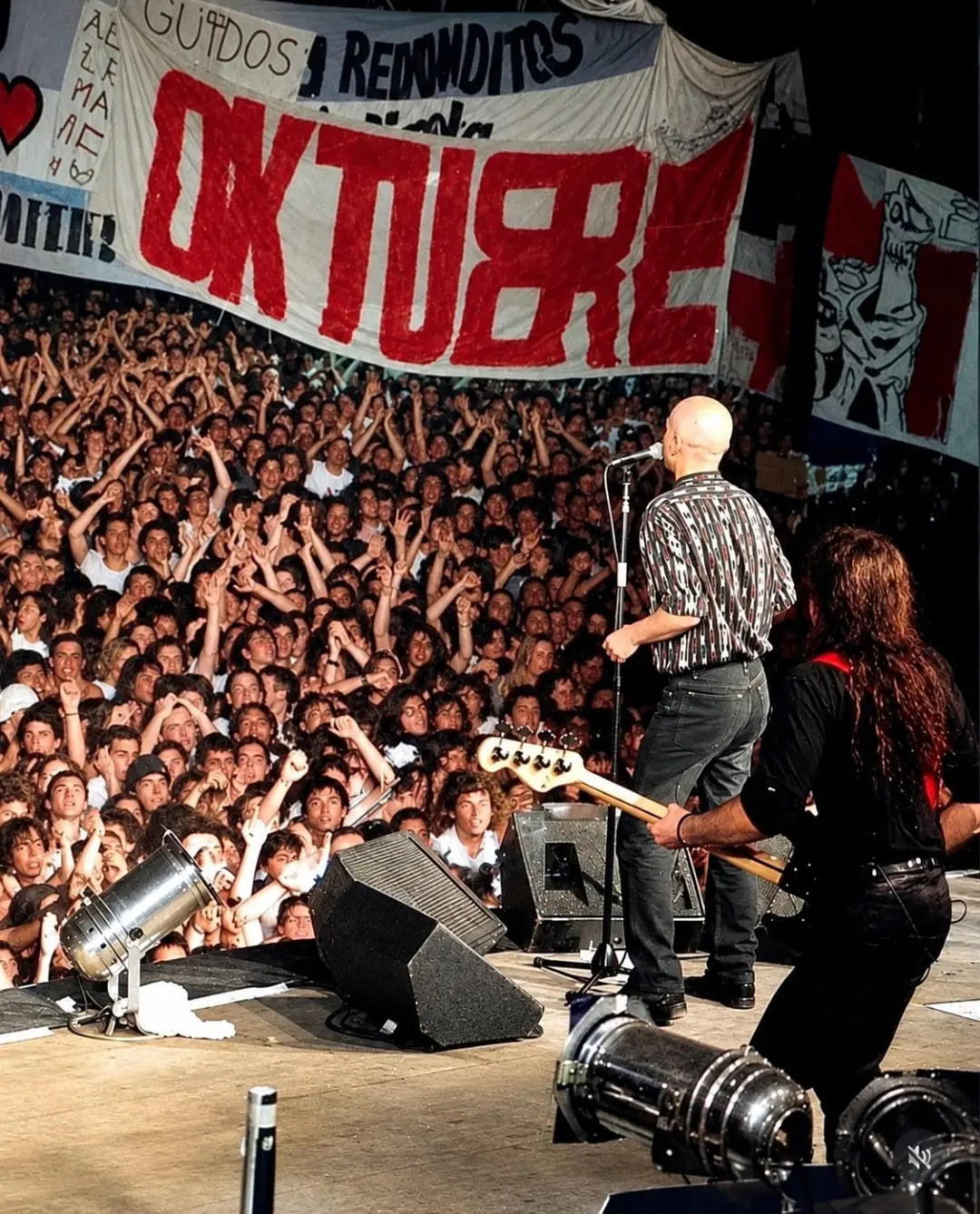 Un cantante pelado canta frente a un micrófono ante una multitud con los brazos en alto y un cartel que dice Oktubre