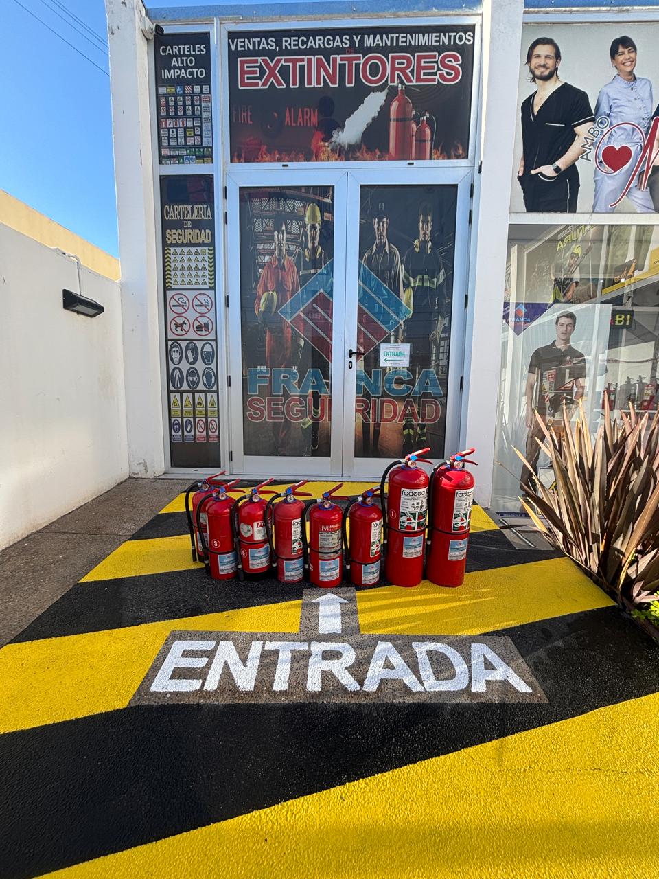 Unos extintores rojos alineados frente a la entrada de un local, sobre un piso amarillo y negro