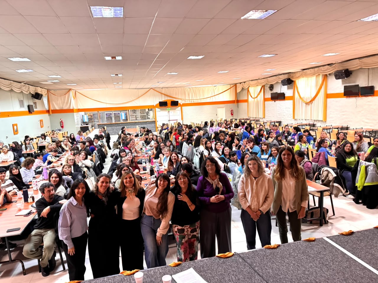 Un grupo de mujeres posa de pie frente a una sala con mesas largas llena de asistentes.