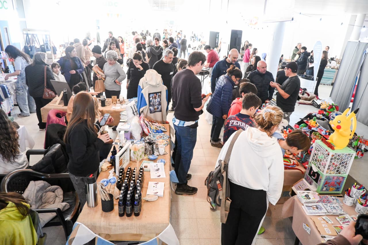 Vista de una feria interior con mesas llenas de productos y personas recorriéndolas
