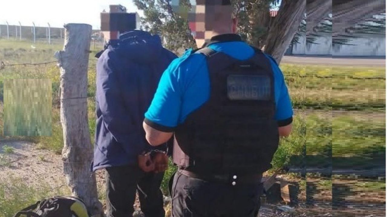 Un policía con chaleco antibalas sujeta de las esposas a un hombre de campera azul junto a un poste de alambrado