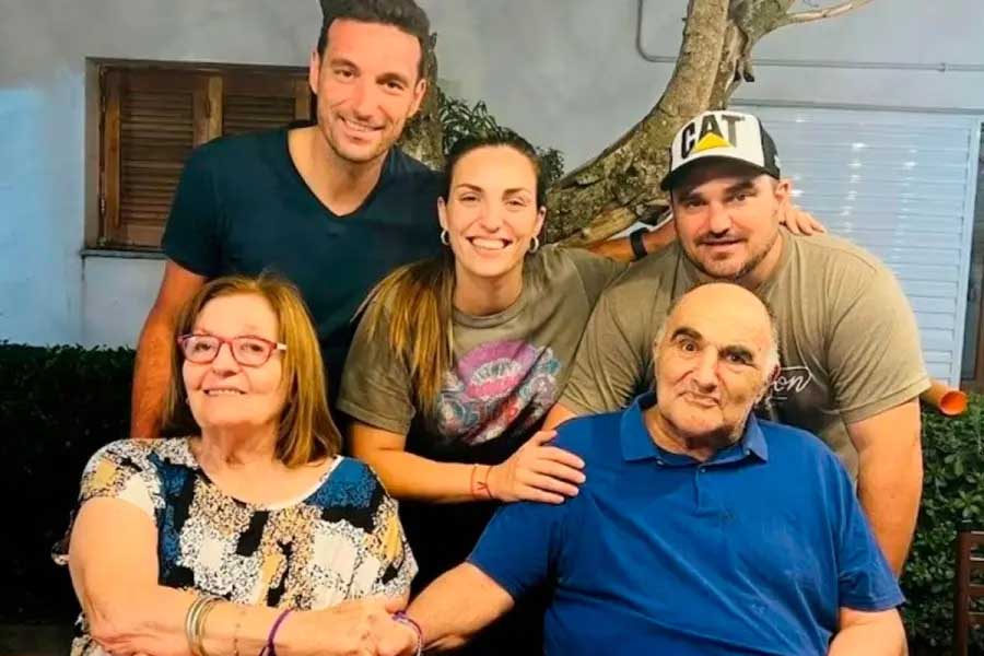 Un grupo de cinco personas posa sonriente frente a una casa; dos están sentadas adelante, una de ellas con anteojos.
