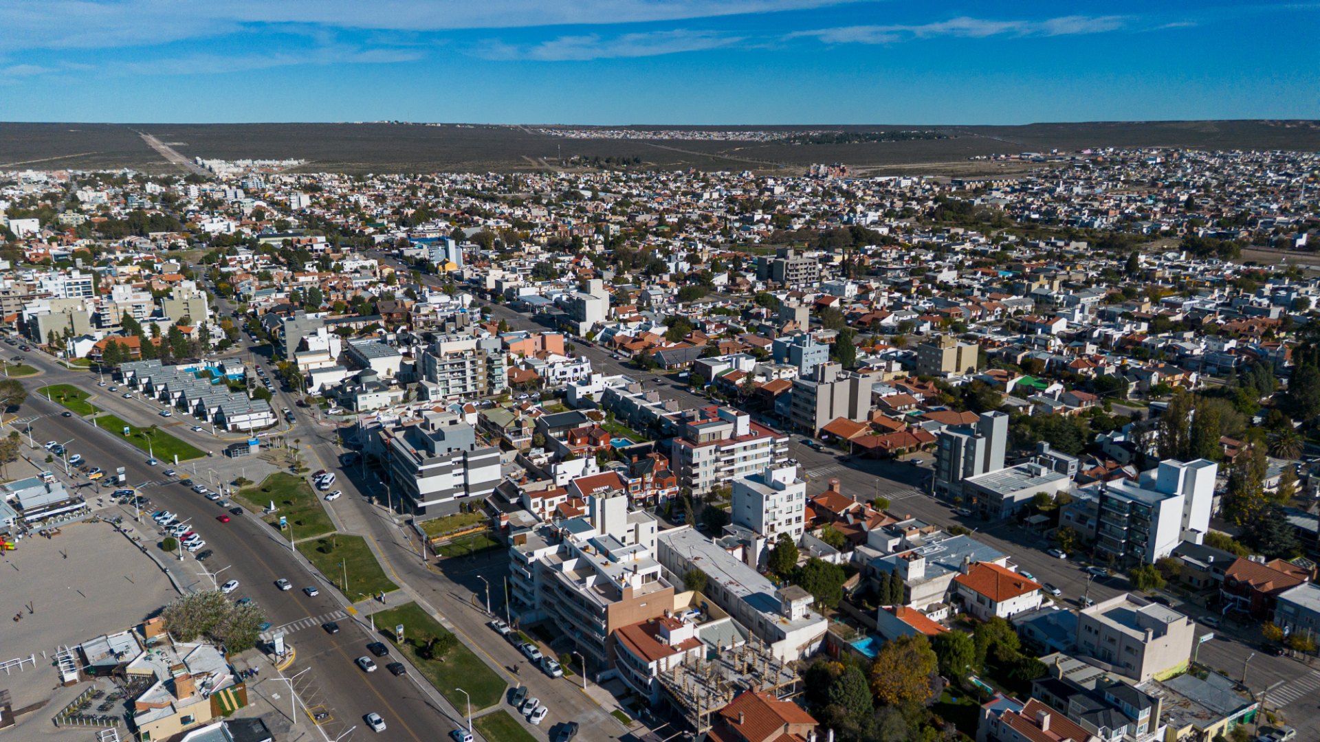 Vista aérea de una ciudad con casas, edificios bajos y una avenida con autos bajo un cielo azul