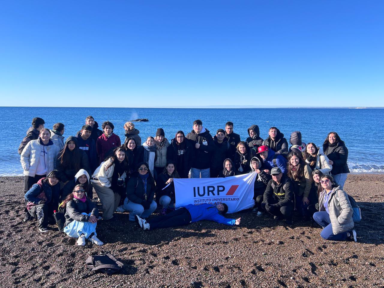 Un grupo de jóvenes posa en una playa de piedra frente al mar con una bandera blanca del IURP