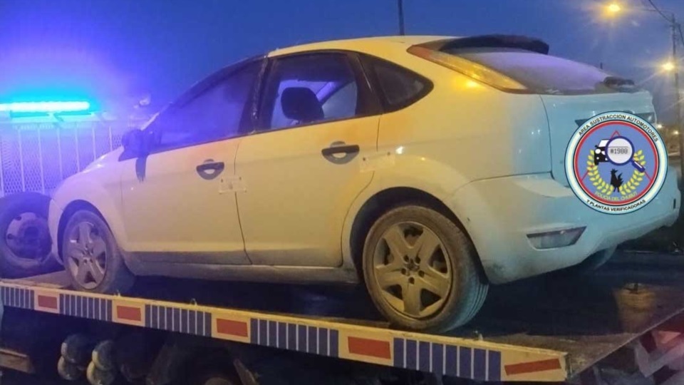 Un auto blanco sobre la plataforma de una grúa, de noche, con luces azules de fondo y una baranda metálica a la izquierda