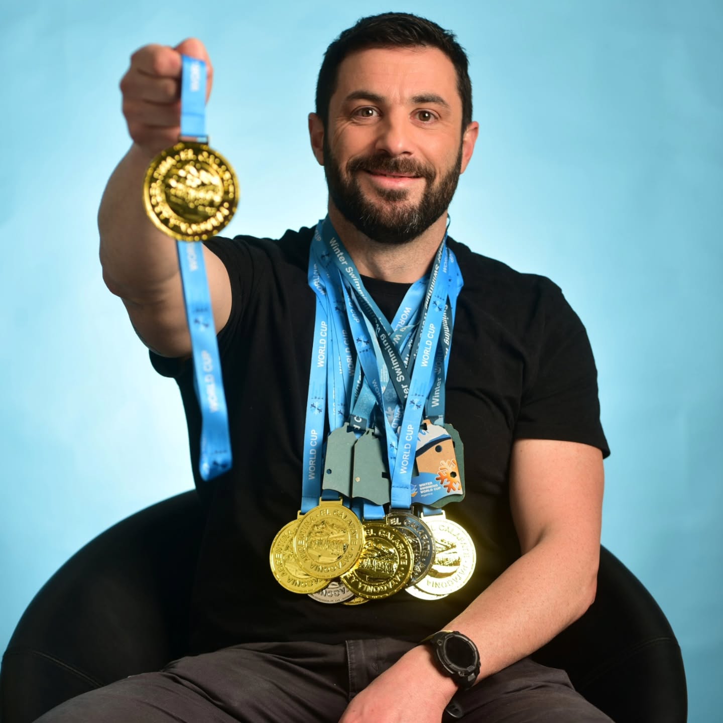 Un hombre sonriente extiende una medalla dorada y lleva varias medallas colgadas al cuello sobre fondo celeste.