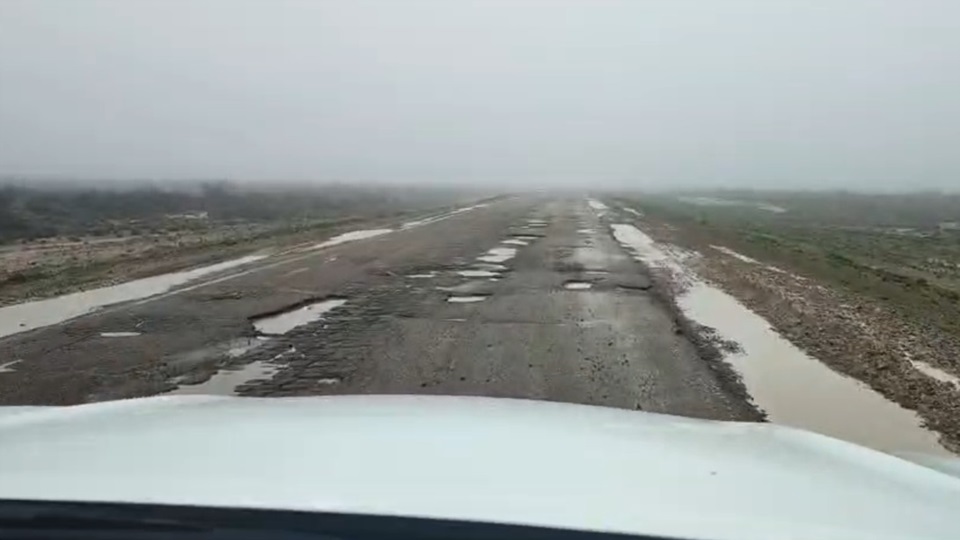 Una ruta con baches y charcos de agua bajo un cielo gris, vista desde el interior de un auto.