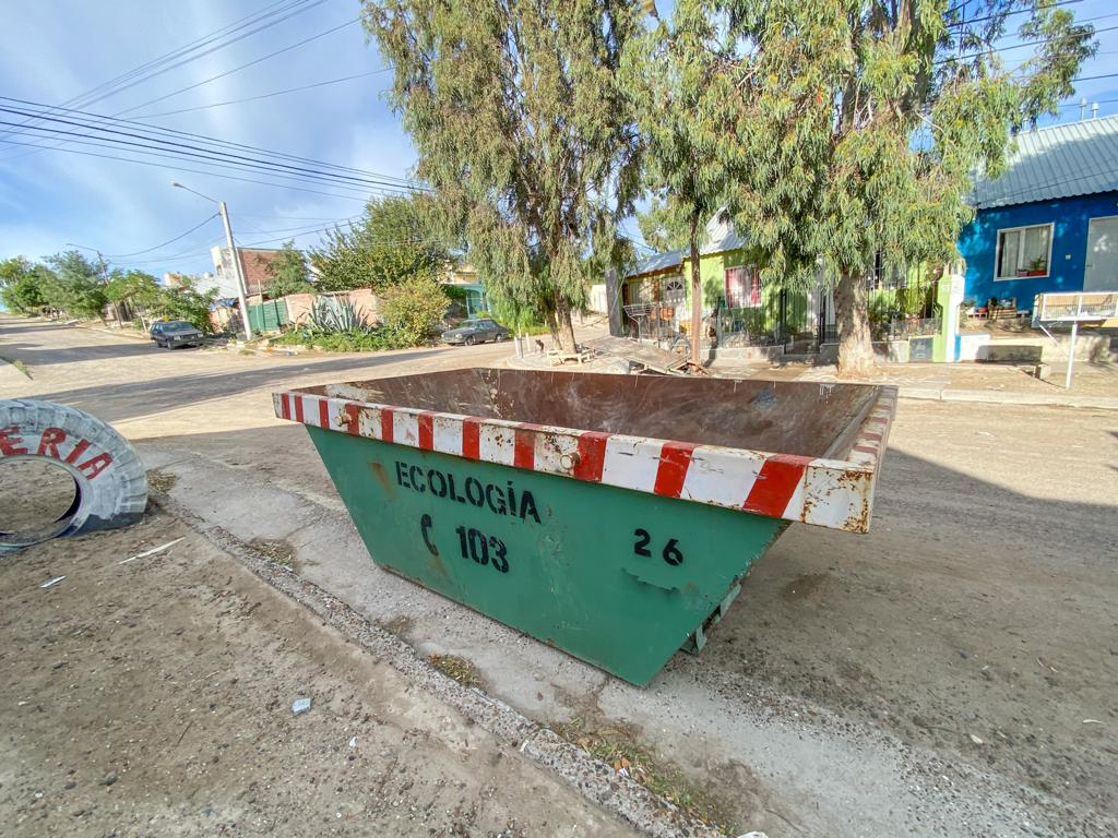 Un contenedor verde y rojo con franjas blancas y la leyenda Ecología sobre una calle de tierra, junto a árboles y casas