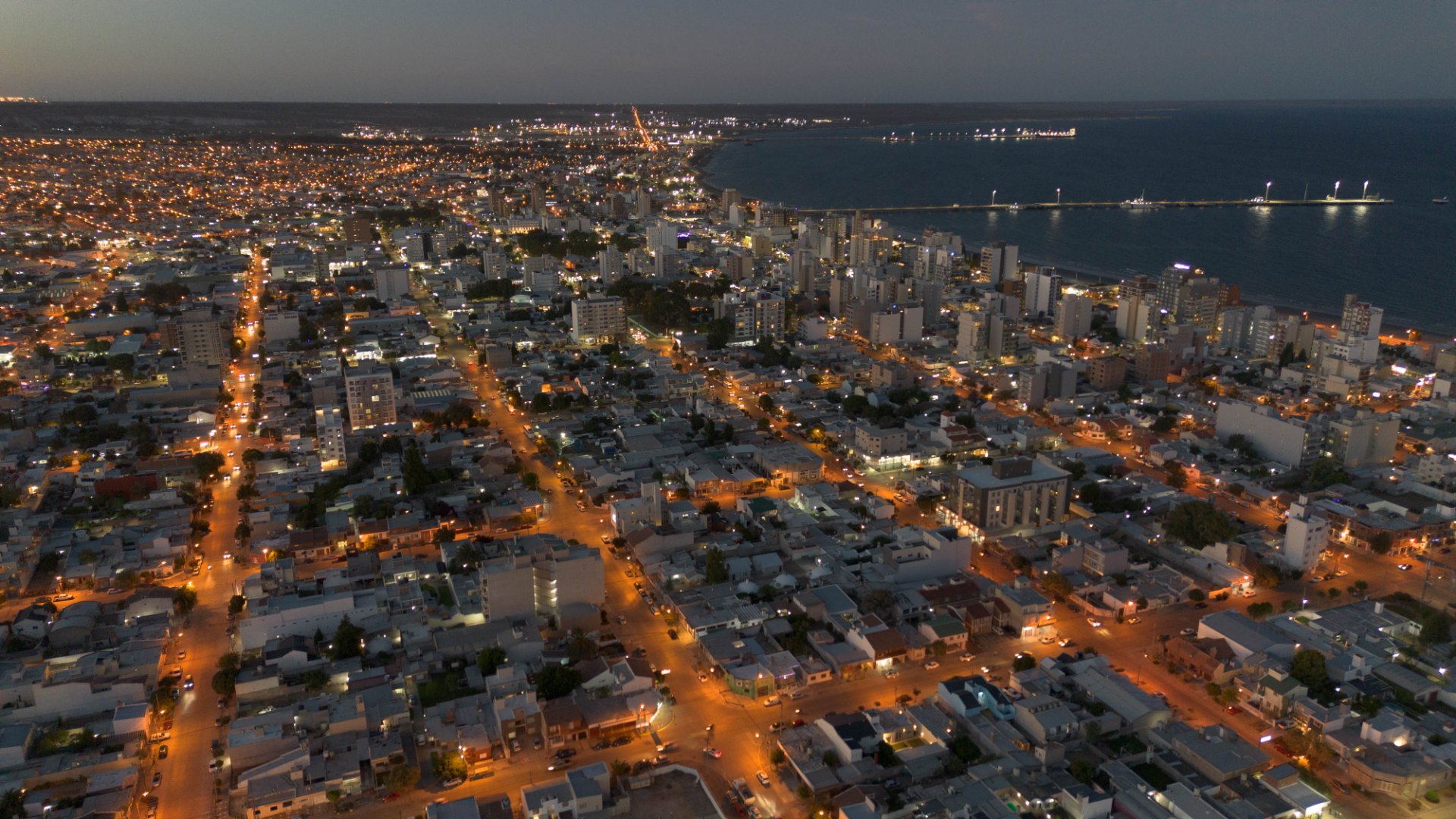 Vista aérea nocturna de la ciudad con calles iluminadas, edificios y el mar al fondo