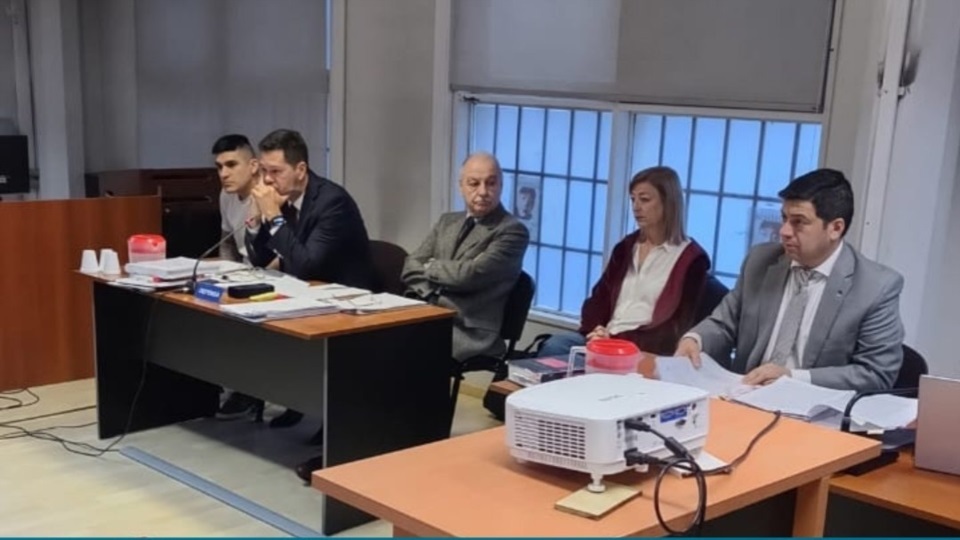 Unas personas sentadas en una sala de audiencias, con una mesa de documentos y un proyector en primer plano.