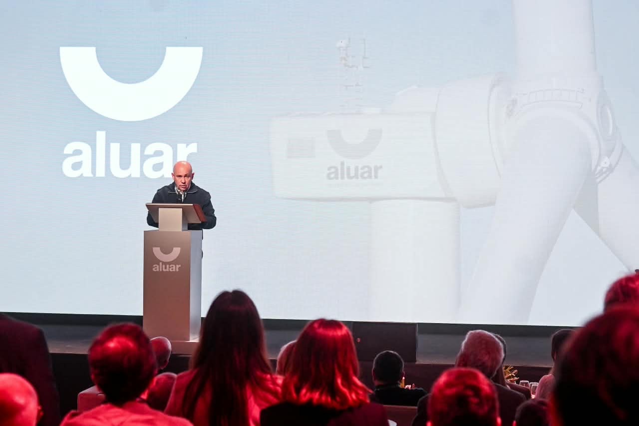 Un hombre habla desde un atril frente a una pantalla con el logo de Aluar y una turbina eólica