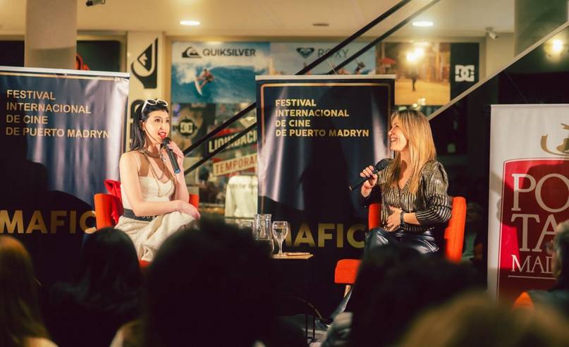 Dos mujeres conversan sentadas en sillones naranjas frente a un público, con micrófonos en la mano