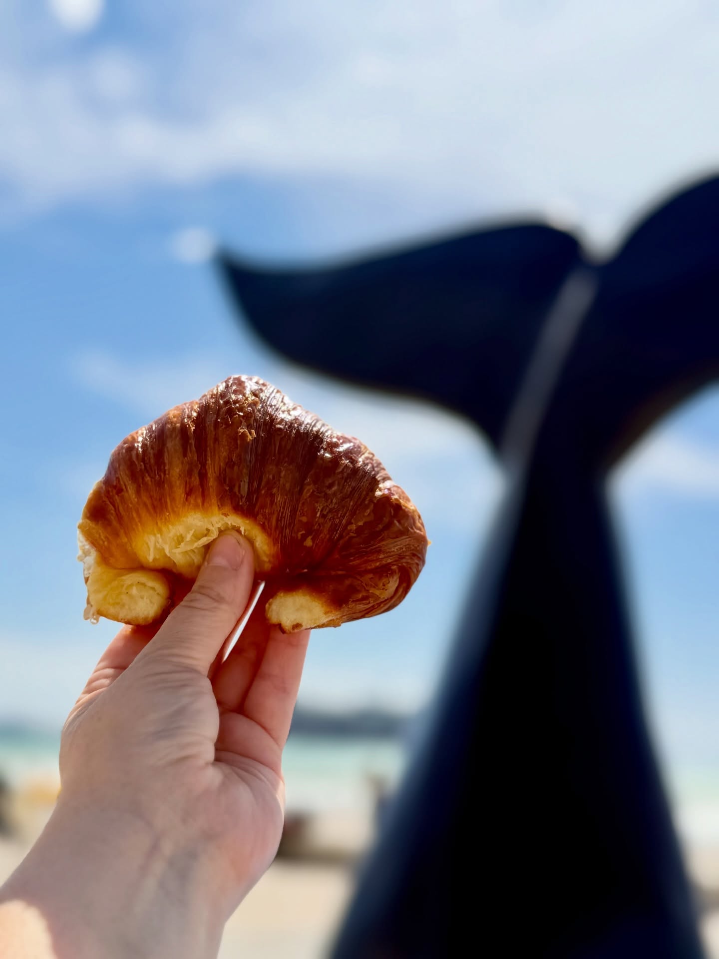 Una mano sostiene un croissant dorado frente al cielo azul, con una cola de ballena oscura desenfocada detrás.